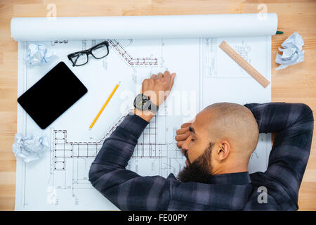 I giovani africani baldheaded americano di giovani designer maschio con la barba giacenti e dormire sul tavolo con blueprint Foto Stock