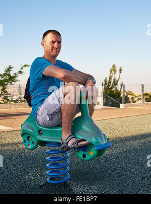 Adulto Uomo bello rivivere l'infanzia giocando in un parco giochi per bambini Equitazione sulla sede della molla. Foto Stock