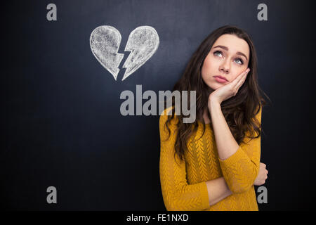 Sconvolto bella giovane donna in piedi e riflettendo su sfondo di lavagna con tratte cuore spezzato Foto Stock