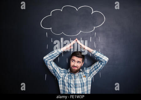 Accigliata irritato giovane tenendo le mani sopra la testa e la copertura dalla pioggia disegnata da raincloud su sfondo blackboard Foto Stock