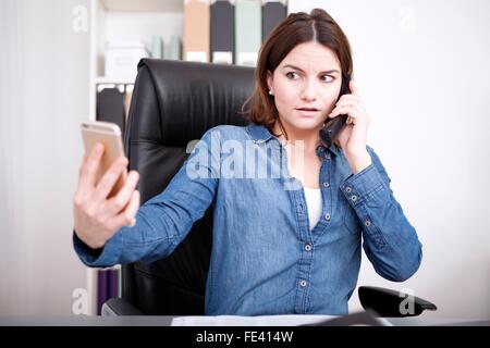 Imprenditrice parlando al telefono di linea terrestre telefono in ufficio e contemporaneamente controllare il suo cellulare con un preoccupato exp Foto Stock