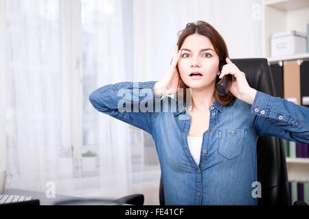 Imprenditrice reagire in stato di shock per una chiamata telefonica seduta alla sua dek con una vasta eyed espressione e la sua mano per il suo volto Foto Stock