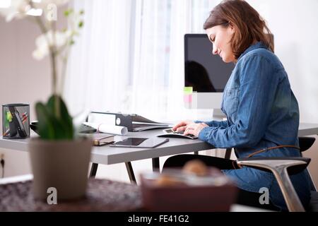 Occupato piuttosto giovane imprenditrice in denim camicetta seduta alla sua tavola portapezzo con documenti e dispositivi elettronici. Foto Stock