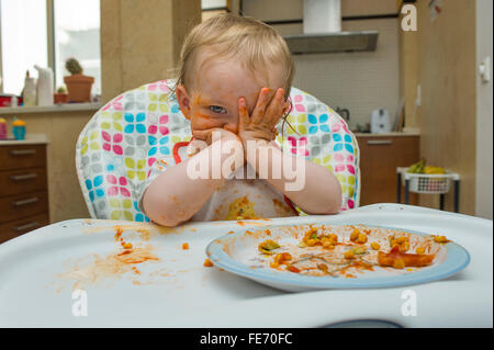 Una bambina (età 15 mesi) mangia un pasto a base di mais dolce e salsa di pomodoro in un modo disordinato e gioca con il suo cibo su una sedia alta Foto Stock