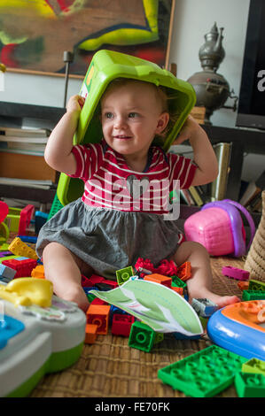 Una bambina (età 15 mesi) è divertente mettere una scatola di plastica sopra la sua testa come lei gioca con Lego e altri giocattoli nel suo soggiorno Foto Stock