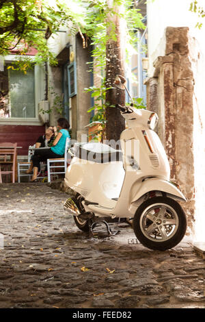 Scooter parcheggiato in acciottolato strada indietro in molivos sull'isola di Lesbo Grecia con due donne di bere il caffè al di fuori di un cafe bar Foto Stock