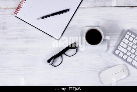 Vista aerea del desktop bianco con parziale tastiera, caffè nero, gli occhiali da lettura, il blocco note e penna e il mouse del computer. Foto Stock