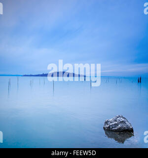 Superficie di acqua, poli, isola e rock in una mattina blu sul lago Trasimeno, Umbria Italia Europa. Colline sullo sfondo. Foto Stock