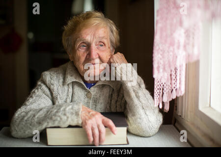 Una donna anziana si siede con un grande libro vicino alla finestra. Foto Stock