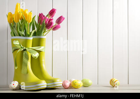Stivali da pioggia, tulipani freschi e uova di pasqua su uno sfondo di legno Foto Stock