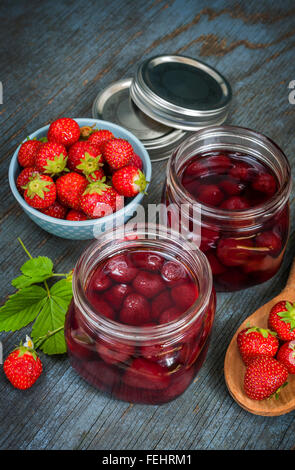 Vasi di vetro con fatti in casa fragola preservare e fragole fresche in un recipiente su blu legno rustico sfondo Foto Stock