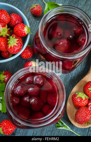 Vasi di vetro con fatti in casa fragola preservare e fragole fresche nel recipiente dal di sopra su blu legno rustico sfondo, close up Foto Stock