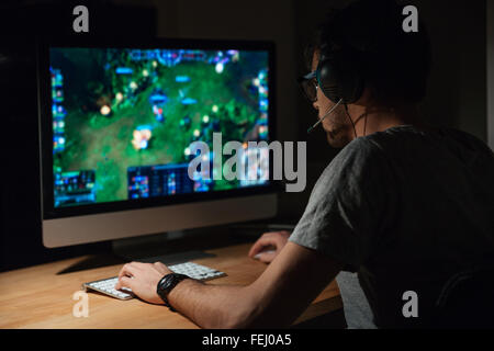 Vista posteriore di concentrato di giovane giocatore in cuffie e occhiali che usano il computer per la riproduzione di gioco a casa Foto Stock