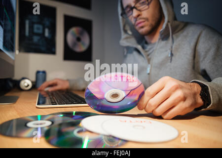 Confuso pensieroso giovane sviluppatore software in felpa con cappuccio e occhiali il lavoro a casa e la scelta di CD con database Foto Stock