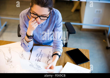 Vista superiore del pensieroso piuttosto giovane donna fashion designer seduto sul posto di lavoro e guardando gli schizzi Foto Stock