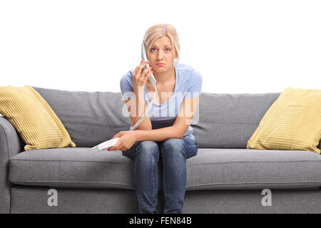 Triste ragazza giovane parlando al telefono seduto su un divano grigio e guardando la telecamera isolata su sfondo bianco Foto Stock