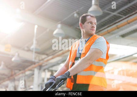 Lavoratore di indumenti da lavoro protettiva tirando il carrello per movimentazione pallet in fabbrica Foto Stock