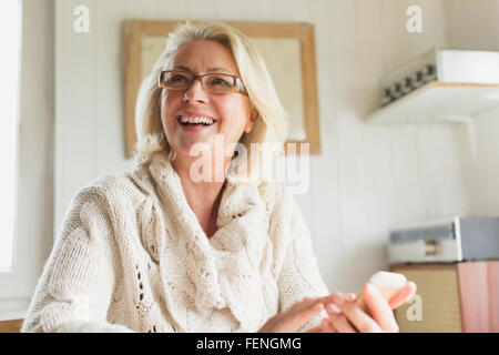 Sorridente donna senior in maglione texting con un telefono cellulare in cucina Foto Stock