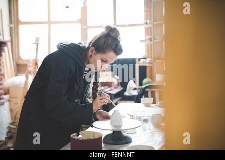 Vista laterale della giovane donna in officina l'applicazione di smalto ceramico alla pentola di creta, guardando verso il basso Foto Stock