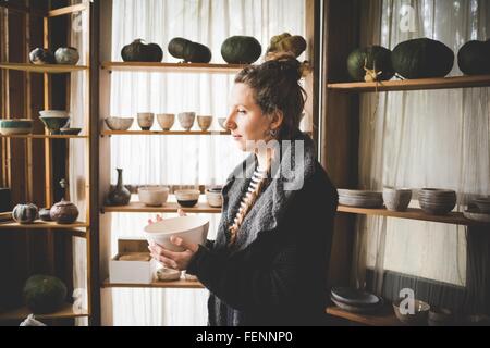 Vista laterale della giovane donna tenendo piatto di ceramica nella parte anteriore degli scaffali la visualizzazione di pentole di creta e zucche Foto Stock