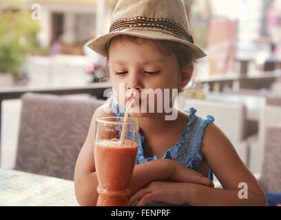 Divertimento kid ragazza in hat frullato di bere succo di vetro in street city cafe Foto Stock