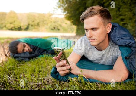 Giovane uomo disteso il sacco a pelo texting sullo smartphone nel campo Foto Stock