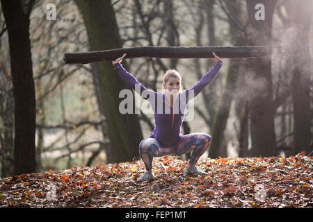 Vista frontale della giovane donna accovacciata nella foresta a braccia alzate tenendo ramo di albero Foto Stock