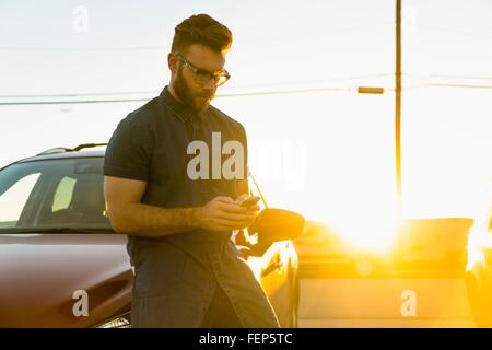 Giovane uomo appoggiato contro auto, utilizza lo smartphone Foto Stock