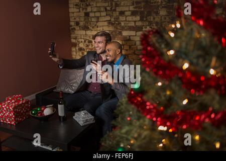 Voce maschile giovane festeggiano il Natale insieme, seduto sul divano, tenendo autoritratto utilizza lo smartphone Foto Stock