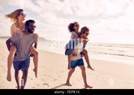 Due belle coppie giovani a piedi dalla spiaggia, con gli uomini che trasportano le loro donne sul loro retro. Le coppie piggybacking sulla riva del mare Foto Stock