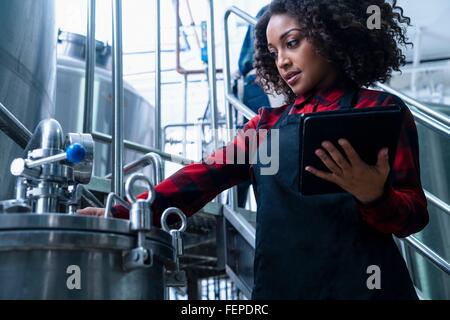 Metà donna adulta nella fabbrica di birra che indossa il grembiule azienda digitale compressa esaminare le attrezzature Foto Stock