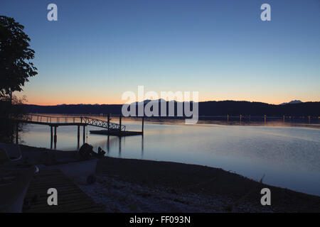 Un molo si estende in un lago calmo, con riflessi della struttura e dell'ambiente circostante riflessi nell'acqua. La scena serena è impreziosita dal tramonto e dal cielo blu, creando un'atmosfera tranquilla all'aperto. Foto Stock