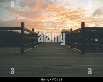 Un molo di legno si estende su un lago sereno, con motivi geometrici nel ponte che conduce verso l'acqua. Il cielo è coperto e le nuvole si riflettono nella superficie ferma del lago, creando un'atmosfera tranquilla. Foto Stock