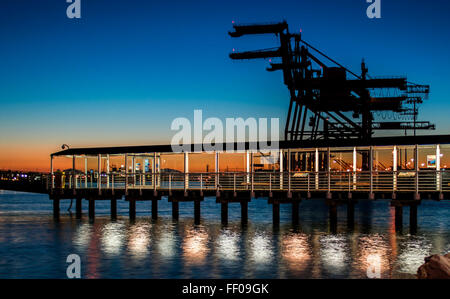 Un molo di legno si estende nelle acque calme al crepuscolo, riflettendo il caldo bagliore della luce del sole che svanisce. La tranquilla scena cattura la quiete della serata, con le luci del molo che proiettano un leggero luccichio sulla superficie dell'acqua. Foto Stock