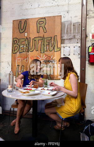 Due donne che condividono un pasto al ristorante Mua a Oakland, in California, l'opera d'arte appesa sul muro dietro di loro legge U R bello. Foto Stock