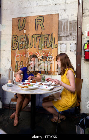 Due donne che condividono un pasto al ristorante Mua a Oakland, in California, l'opera d'arte appesa sul muro dietro di loro legge U R bello. Foto Stock