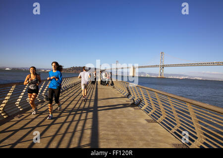 Persone fare jogging lungo il San Francisco Pier 14 in un pomeriggio soleggiato con il ponte della baia in background. Foto Stock