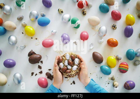 Le mani del bambino tenendo piccolo nido o cesto con le uova di Pasqua di cioccolata oltre quelle dipinte Foto Stock