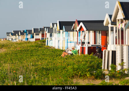 Donna seduta sulla sedia a sdraio e la lettura di giornale in legno colorato cambiando case sulla spiaggia di Skanör nella Svezia meridionale Foto Stock