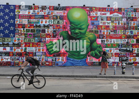 Un uomo di cicli di lavoro passato un Ron English art collage su Bowery murale sulla parete Houston Street a New York Foto Stock