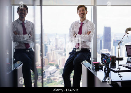 Imprenditore adulto seduto in un ufficio moderno con una bella vista della città. L'uomo guarda la telecamera e sorrisi con bracci cr Foto Stock