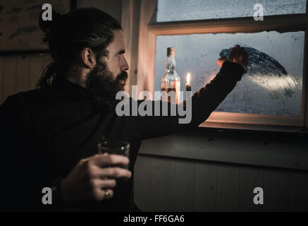 Un uomo seduto da solo in una stanza con una bottiglia di whisky e un bicchiere, tergi condensazione finestra off Foto Stock