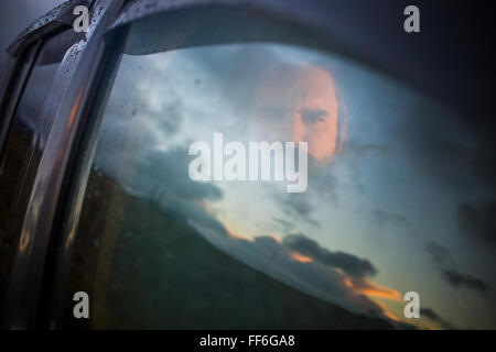 Un uomo seduto in un auto guardando fuori. Riflessioni del Cielo di tramonto sulla finestra. Foto Stock
