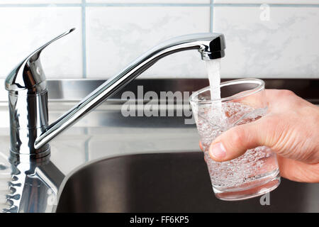 Mano che tiene un bicchiere di acqua versata dal rubinetto di cucina. Foto Stock