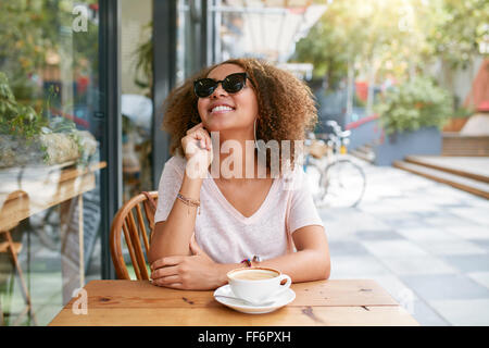 Ritratto di giovane donna seduta al cafè sul marciapiede cercando felice. Femmina africana udienza presso la caffetteria con cappuccino. Foto Stock