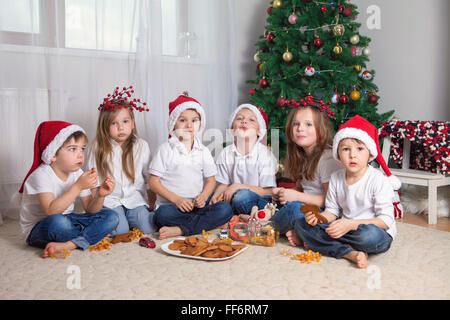 Sei adorabili bambini divertirsi nella parte anteriore dell'albero di Natale, mangiando caramelle e biscotti Foto Stock