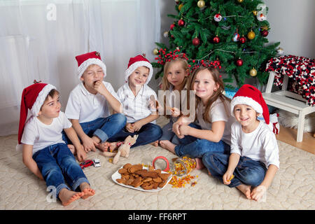 Sei adorabili bambini divertirsi nella parte anteriore dell'albero di Natale, mangiando caramelle e biscotti Foto Stock