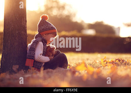 Adorable little boy con Teddy bear nel parco in una giornata autunnale nel pomeriggio, seduto sull'erba Foto Stock