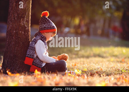 Adorable little boy con Teddy bear nel parco in una giornata autunnale nel pomeriggio, seduto sull'erba Foto Stock