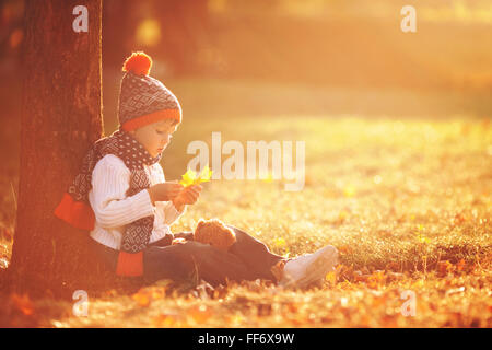 Adorable little boy con Teddy bear nel parco in una giornata autunnale nel pomeriggio, seduto sull'erba Foto Stock